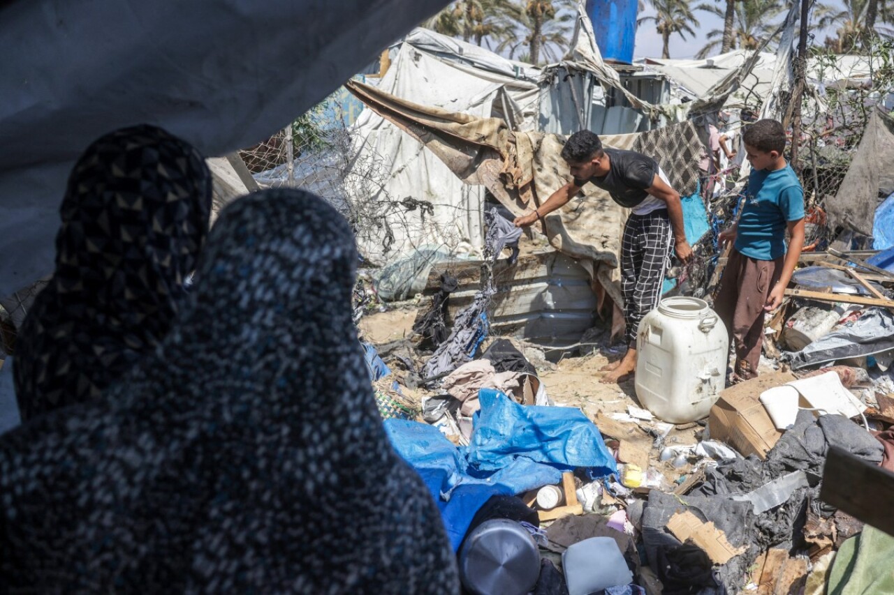 A woman in black clothing watches from the foreground as several children and adults move about a sandy area with makeshift shelters, scattered belongings, and debris.
