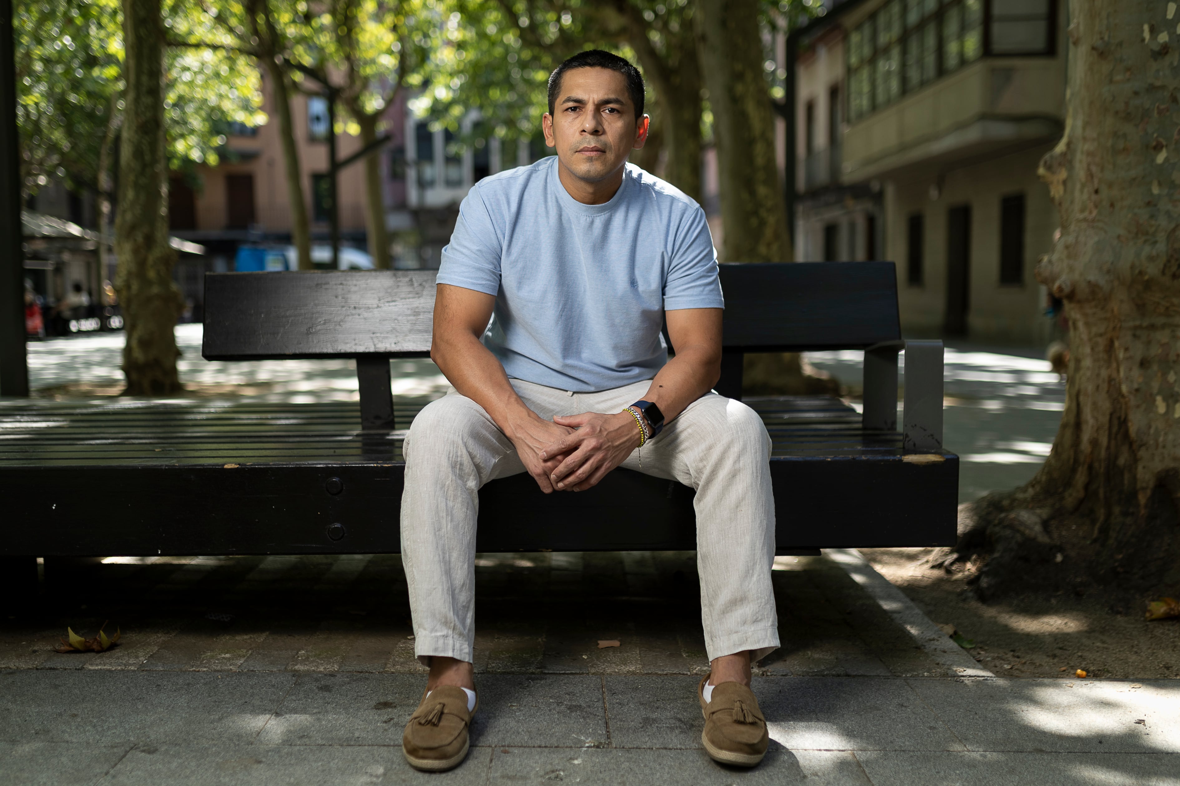 A young man sitting on a wooden bench in a tree-lined plaza in an urban neighbourhood, looking at the camera with a serious expression.