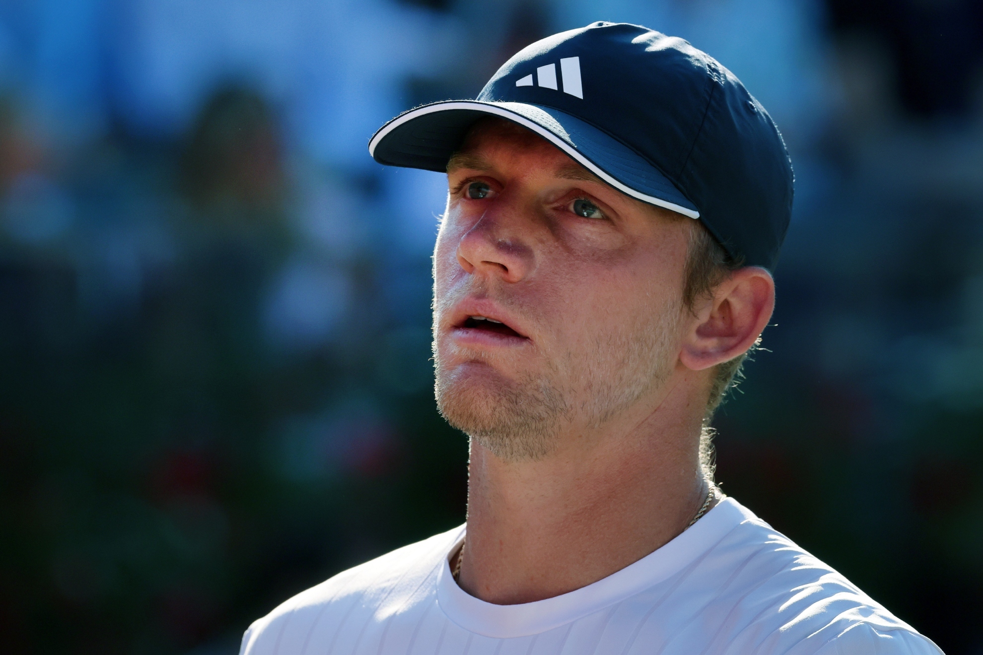 A tennis player in an Adidas blue cap and white shirt looks upward in sunlight.
