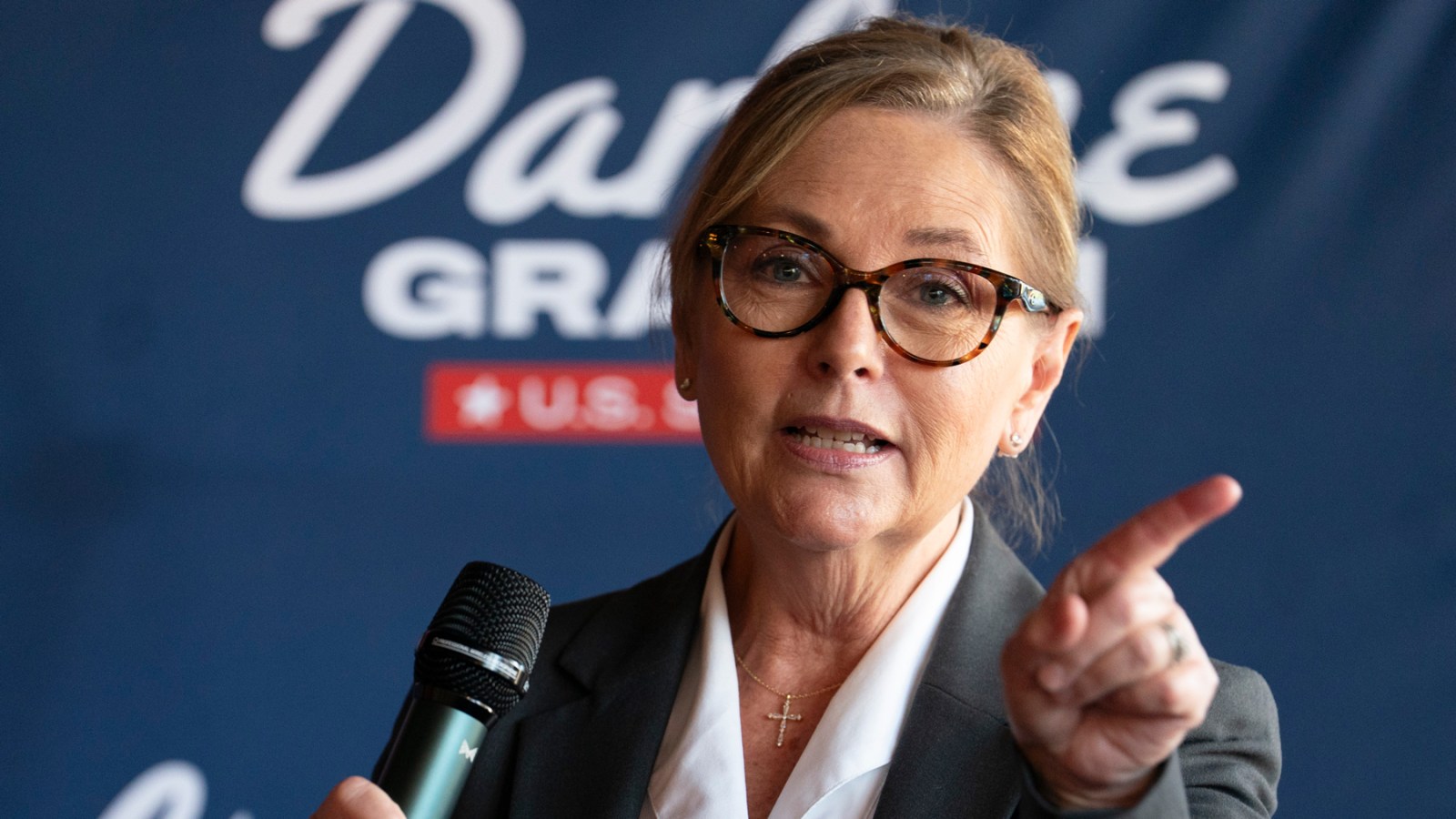 A woman wearing glasses and a gray blazer speaks into a microphone in front of a blue backdrop with partially visible text.
