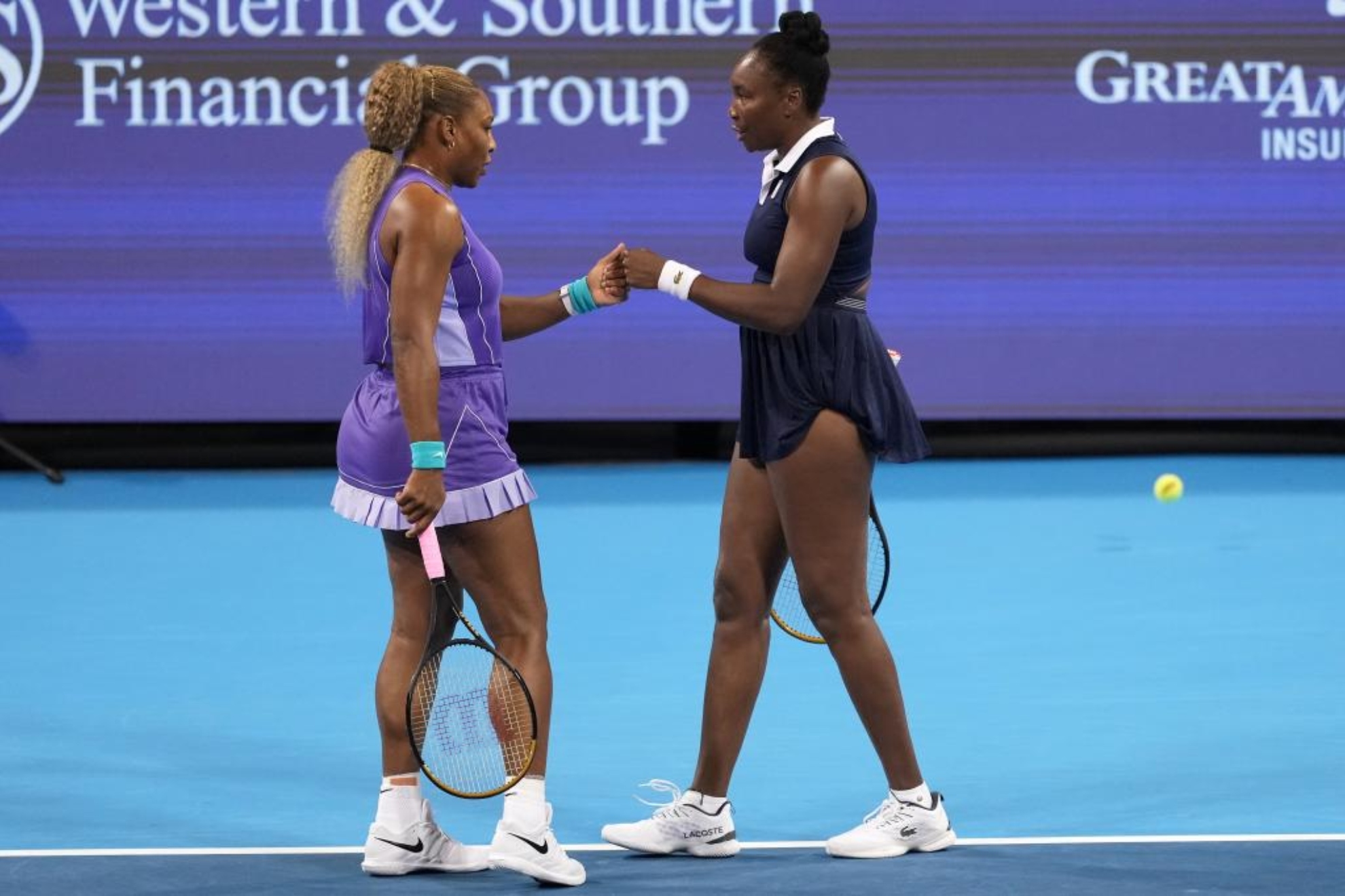 Two female tennis players on a blue hard court embrace and touch fists during a match, with sponsor logos visible on the backdrop.