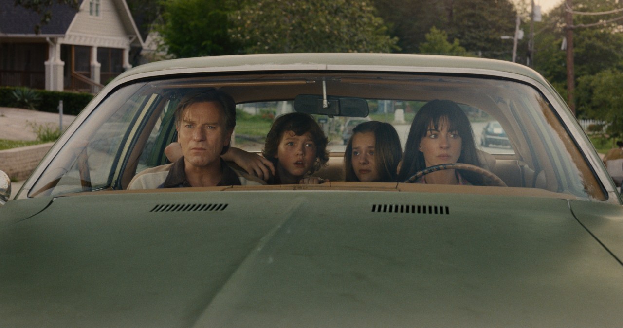 Four people sit in the front seat of a vintage car, looking ahead with concerned expressions on a residential street lined with houses.