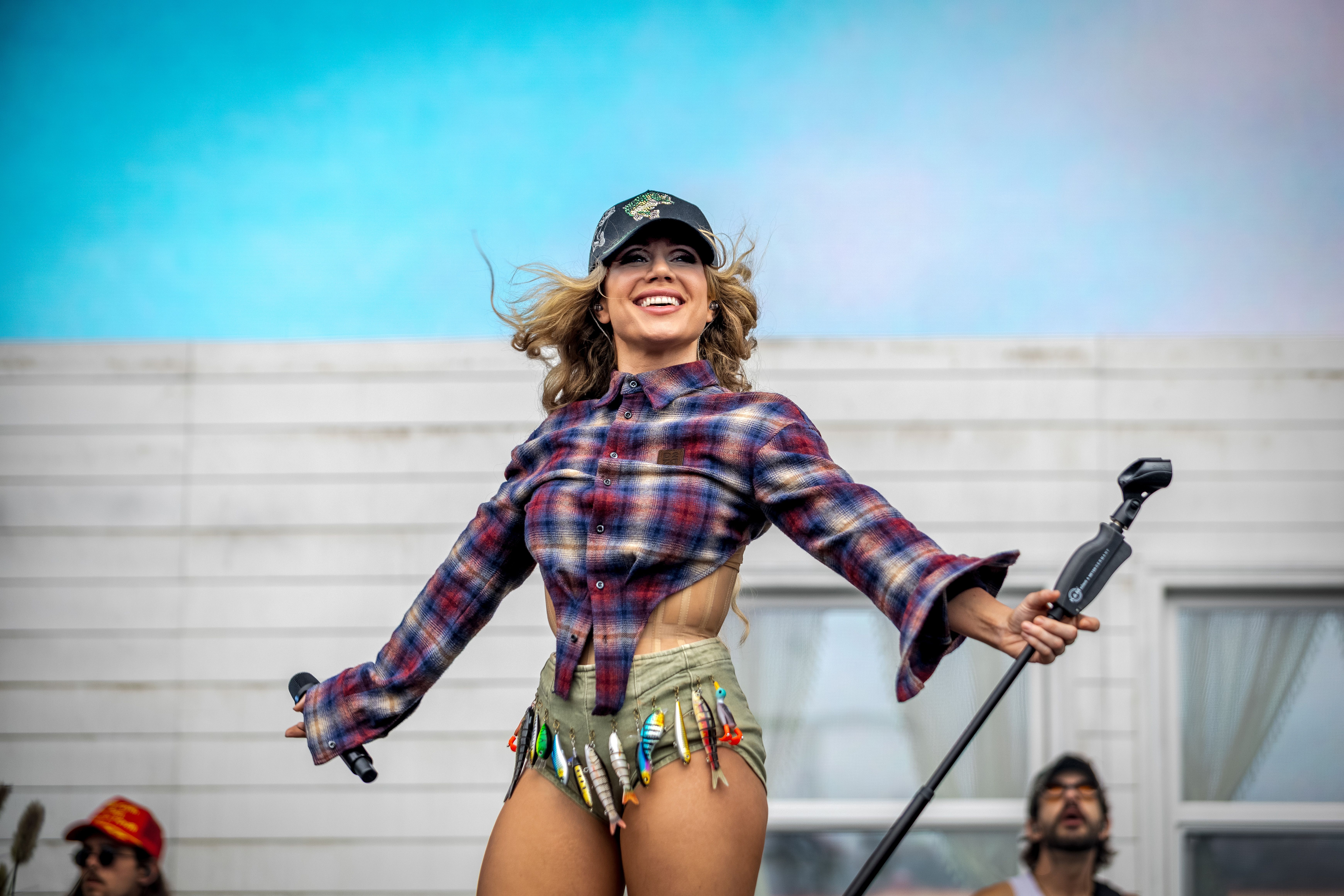 A performer on stage wearing a cropped plaid shirt, khaki shorts with fishing lures hanging from the waistband, a black cap, and holding a microphone stand, smiling at the audience under a blue sky with a white building in the background.