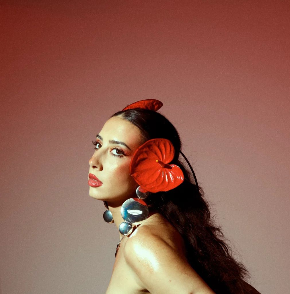 A woman in profile against a mauve background wears red petal-shaped ornaments in her hair and matching red drop earrings with turquoise stones.