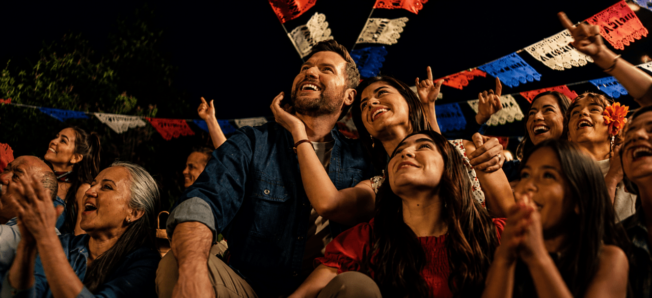Un grupo de personas de múltiples generaciones, incluyendo adultos mayores, adultos jóvenes y niños, celebran juntos bajo decoraciones de papel picado rojo, blanco y azul iluminadas en la noche.