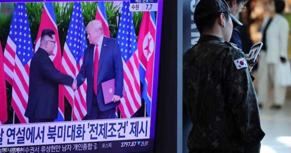 A television screen shows two men in dark suits shaking hands in front of U.S. and North Korean flags, as people watch the broadcast at a train station.