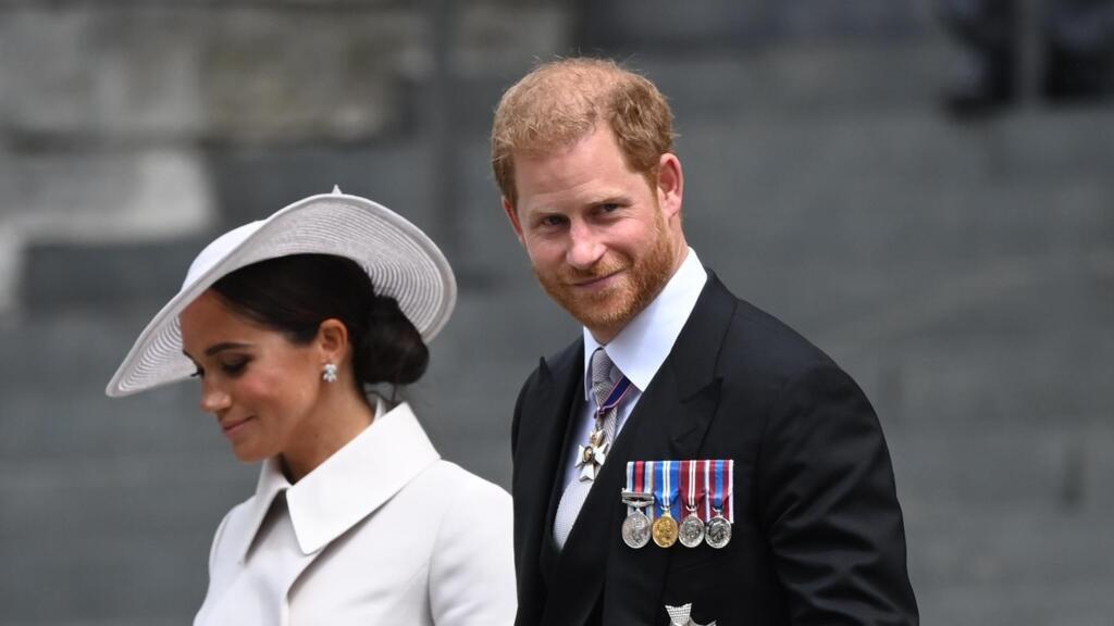Un hombre con uniforme militar negro con medallas y una mujer con un traje blanco y sombrero de ala ancha caminan juntos en un exterior urbano.