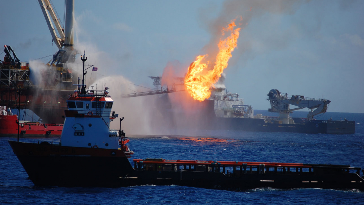 A large flame shoots from an offshore oil platform in the ocean, with a supply vessel and fire suppression equipment nearby.