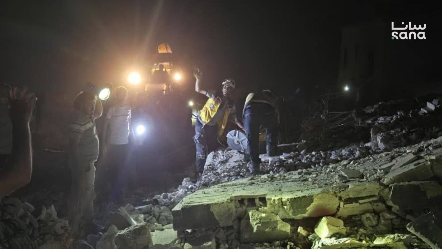 Rescue workers work at night among rubble and debris of a destroyed building, illuminated by torches and floodlights.
