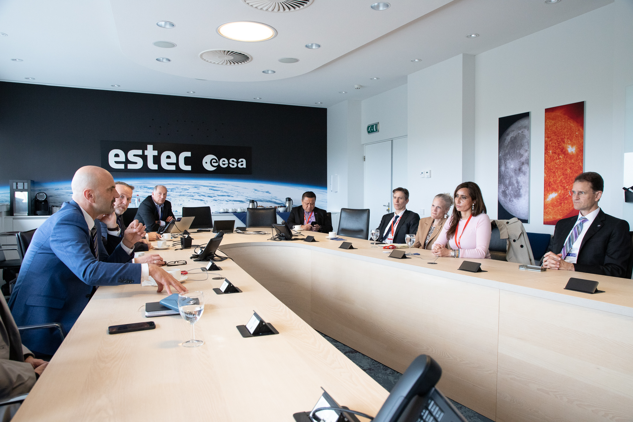 Un grupo de profesionales en traje de negocios se reúne alrededor de una larga mesa de conferencias en una sala de reuniones moderna con el logo de "estec @esa" en la pared de fondo.