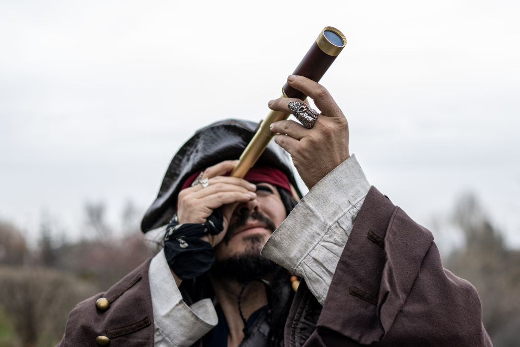 Un hombre viste un traje de época estilo pirata con tricornio negro, levantando un telescopio hacia el cielo mientras porta anillos decorativos en sus manos.