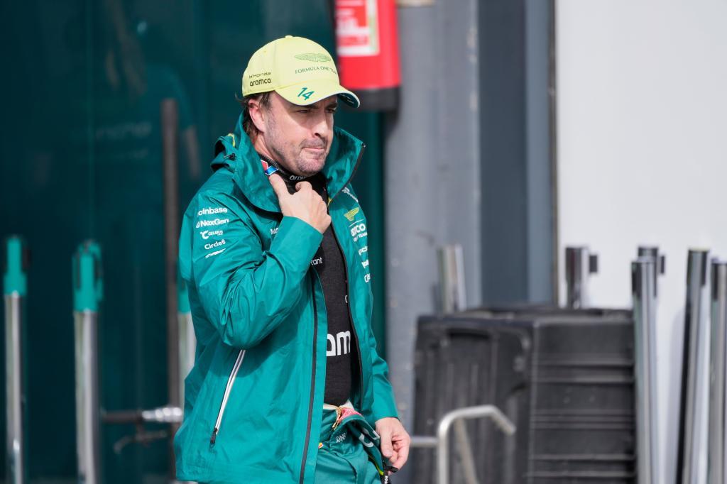 A racing driver in a turquoise jacket and yellow cap walks through a paddock area, adjusting his helmet.