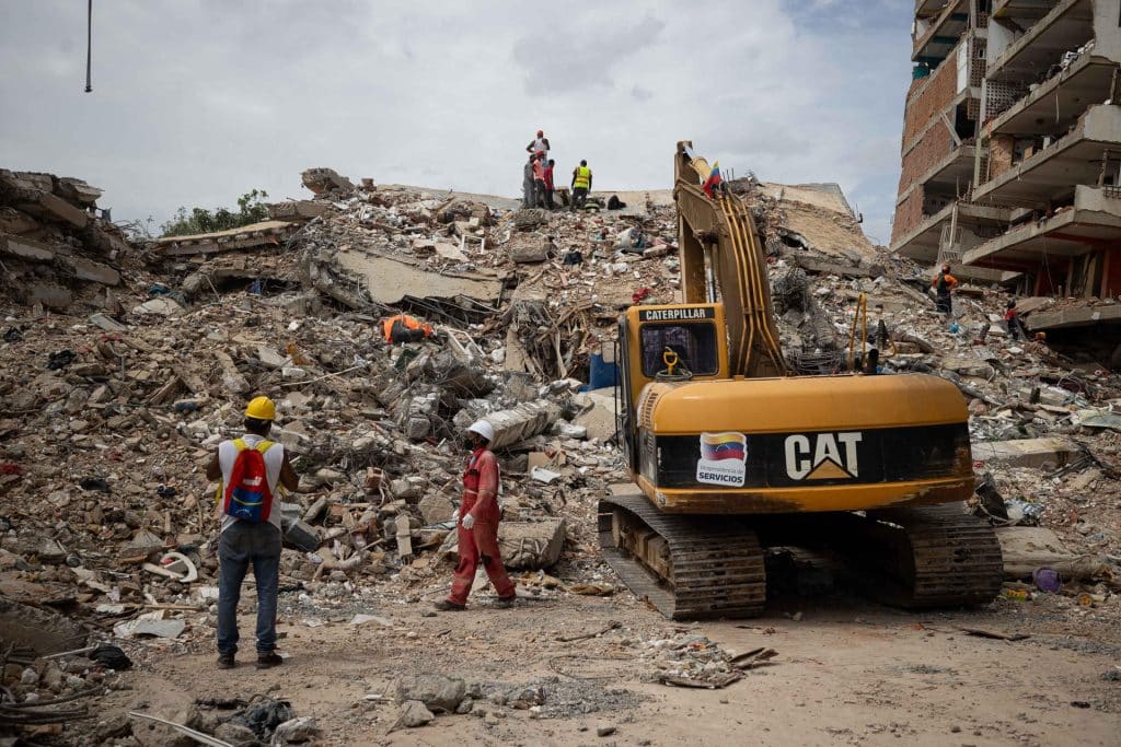 A Caterpillar excavator removes debris from destroyed buildings in an urban area, while workers wearing hard hats and safety vests supervise cleaning operations.