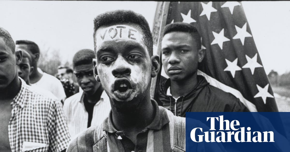 A black and white photograph of a protest where a young man with the word "VOTE" written on his forehead holds an American flag, surrounded by other participants.