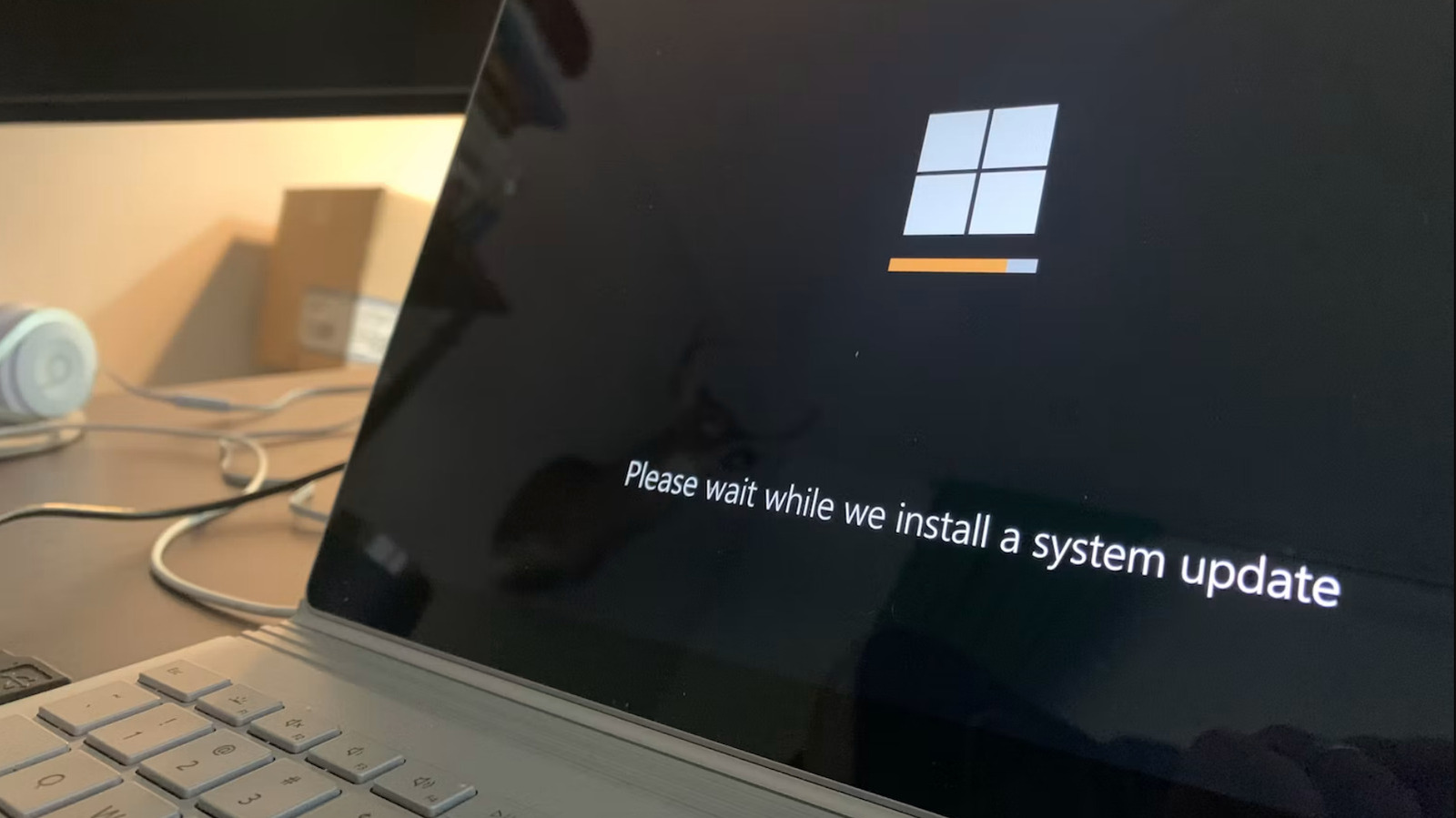 Un portátil mostrando la pantalla de instalación de Windows con el logotipo de Windows, una barra de progreso naranja y el mensaje "Please wait while we install a system update".