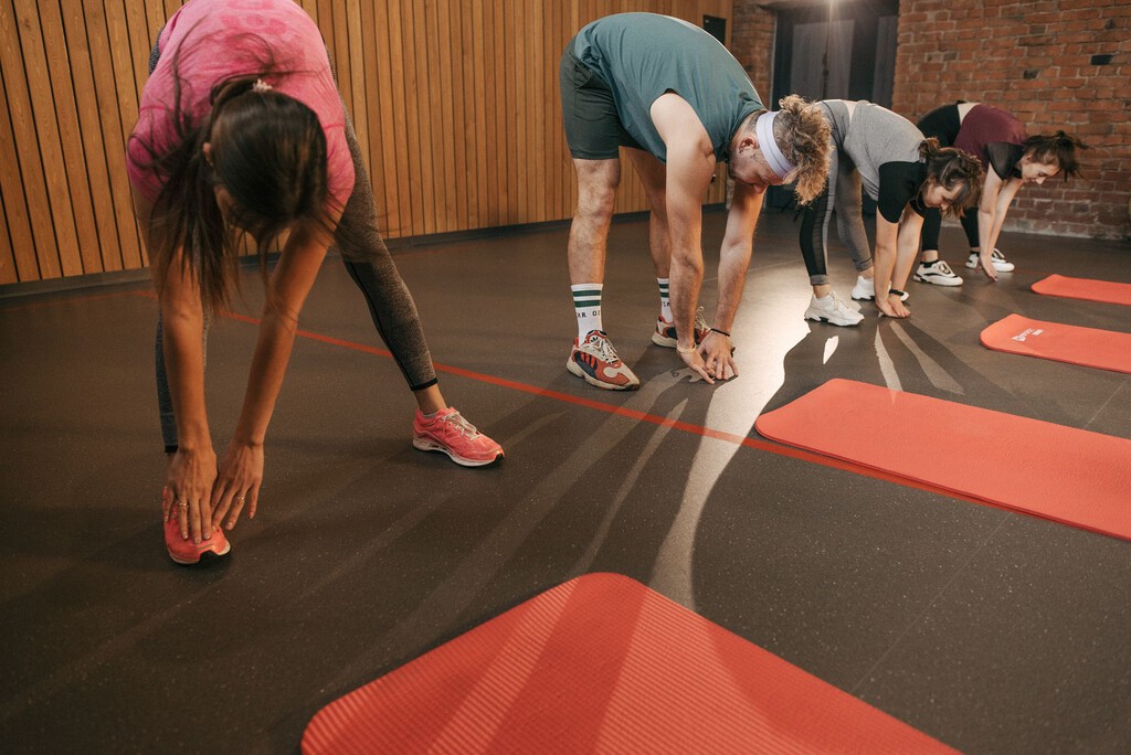 Un grupo de personas de diferentes edades realiza ejercicios de flexibilidad y calentamiento en una sala de fitness moderna con piso de caucho negro y bandas de colores naranja, inclinadas hacia adelante con las manos extendidas hacia el piso.