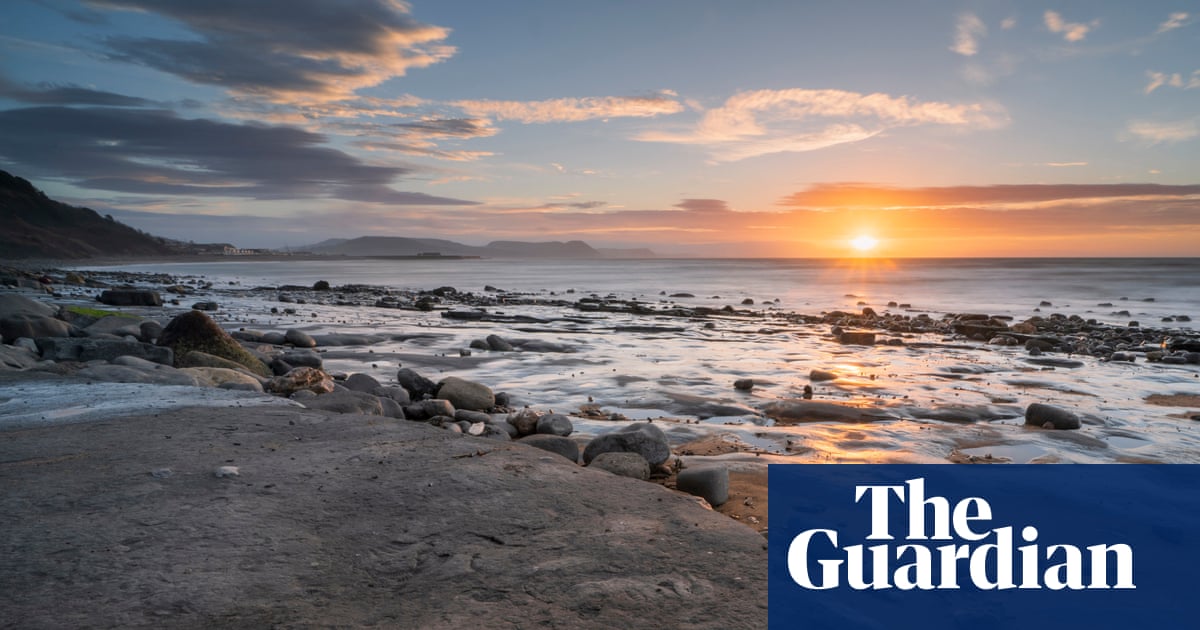 A rocky beach at sunset with waves lapping against boulders, mountains visible across the water, and dramatic clouds illuminated by orange and golden light on the horizon.