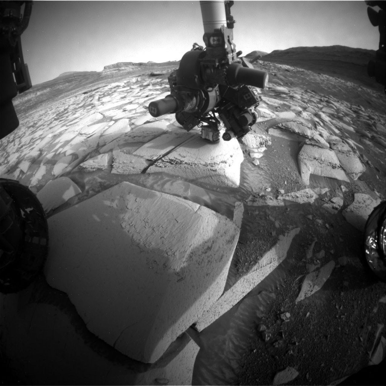 A robotic rover with articulated arms and instruments examines layered rock formations on a dusty, barren landscape with hills in the background.