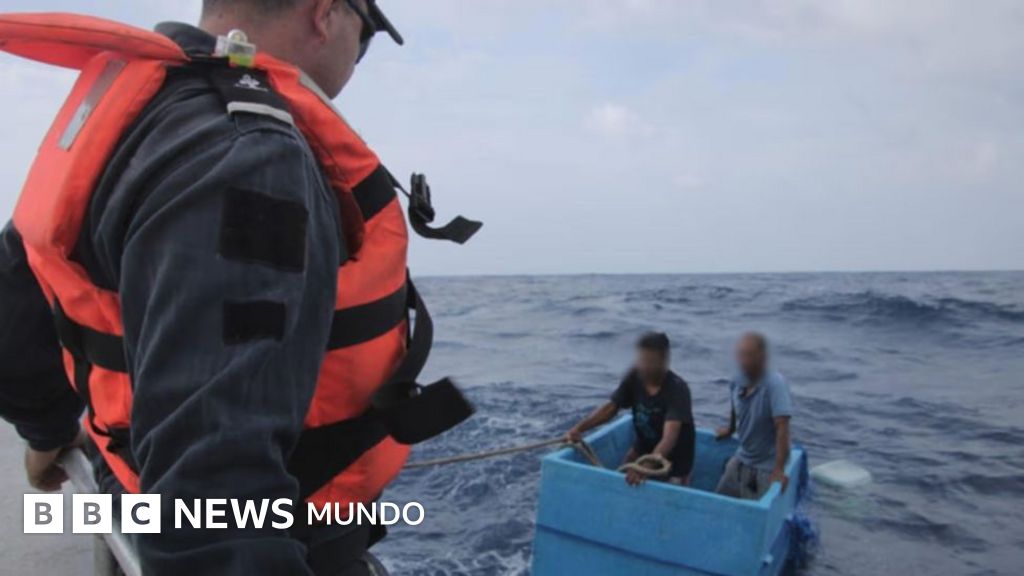 Un rescatista con chaleco salvavidas rojo ayuda a dos hombres en una pequeña embarcación azul en el océano.