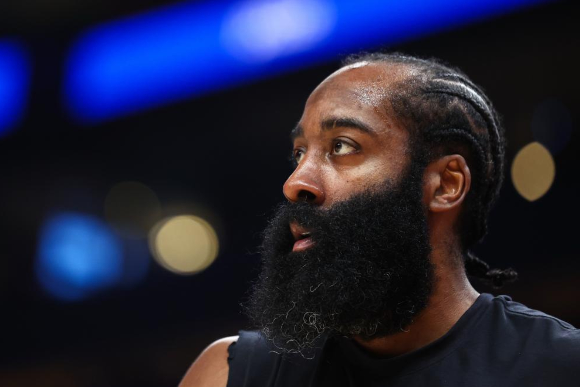 A basketball player seen in profile during a game, with distinctive beard, wearing a dark sleeveless uniform against a blurred background with blue lighting.