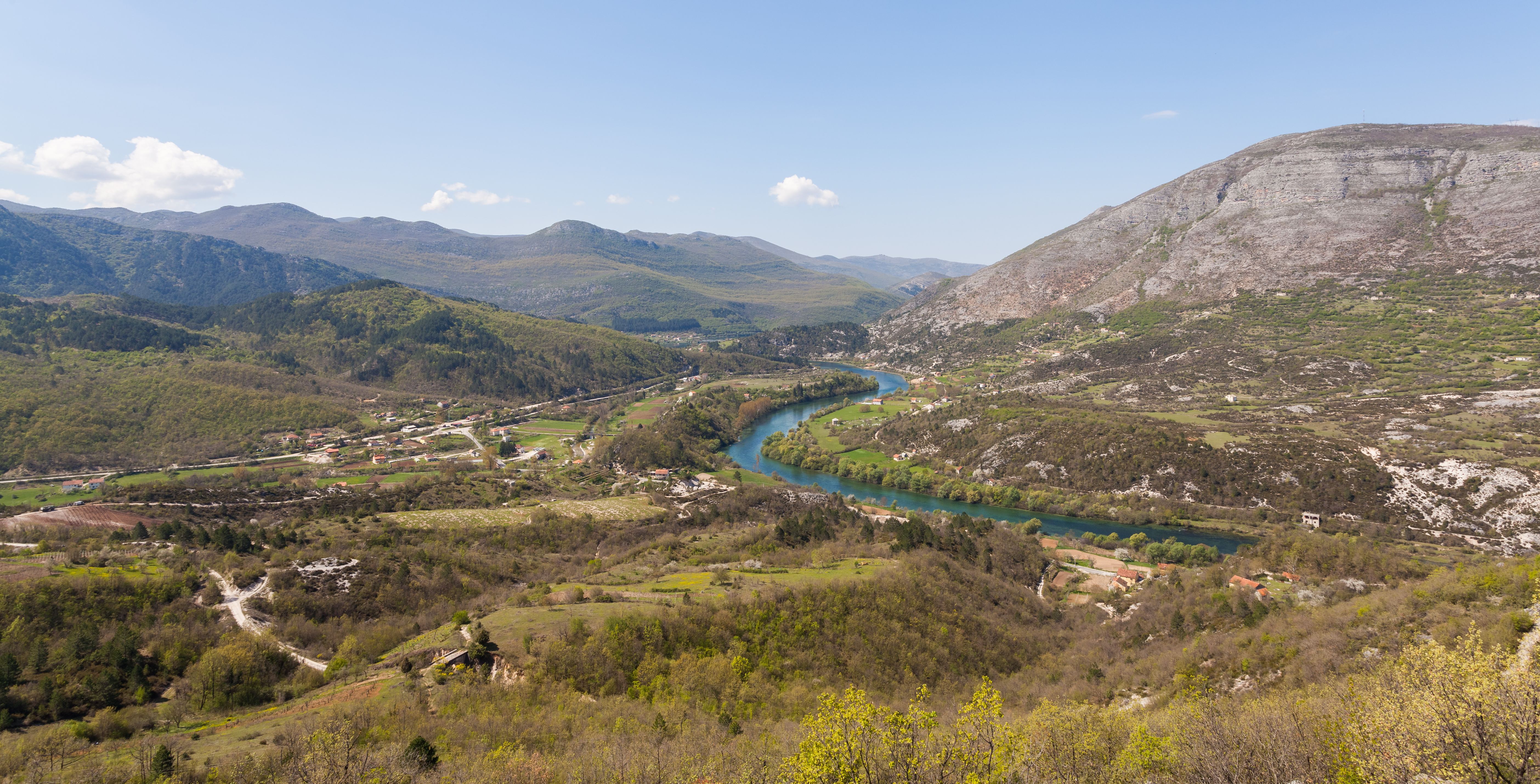 An aerial view of a river valley surrounded by limestone mountains and dense green forests, with a bright blue-green river winding through the landscape.