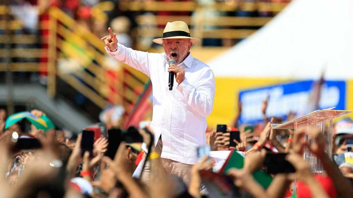 Un hombre de edad avanzada con sombrero de paja y camisa blanca sostiene un micrófono mientras gesticula ante una multitud, con gradas y espectadores borrosos al fondo.