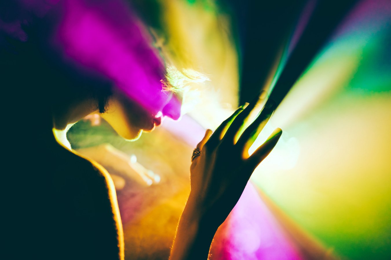 A person with hands outstretched is illuminated by vibrant coloured lights including magenta, yellow, green and orange in a dark environment.