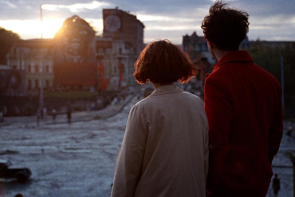 Dos personas de espaldas observan una ciudad al atardecer, con edificios y el sol poniéndose al fondo.