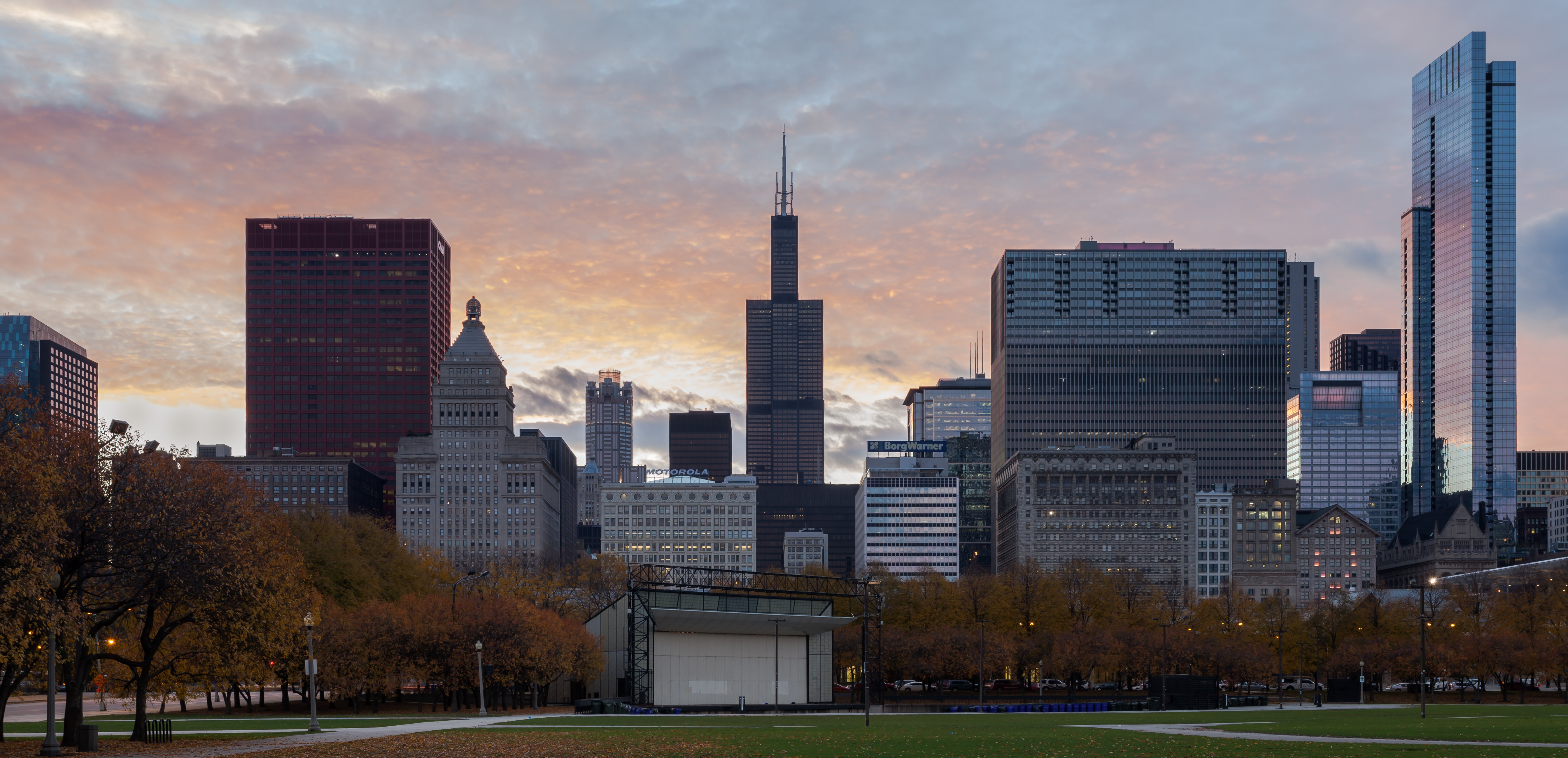 Chicago's downtown skyline at sunset with tall buildings, including a distinctive dark skyscraper with antenna spires in the center, overlooking Grant Park with bare autumn trees and a stage structure in the foreground.