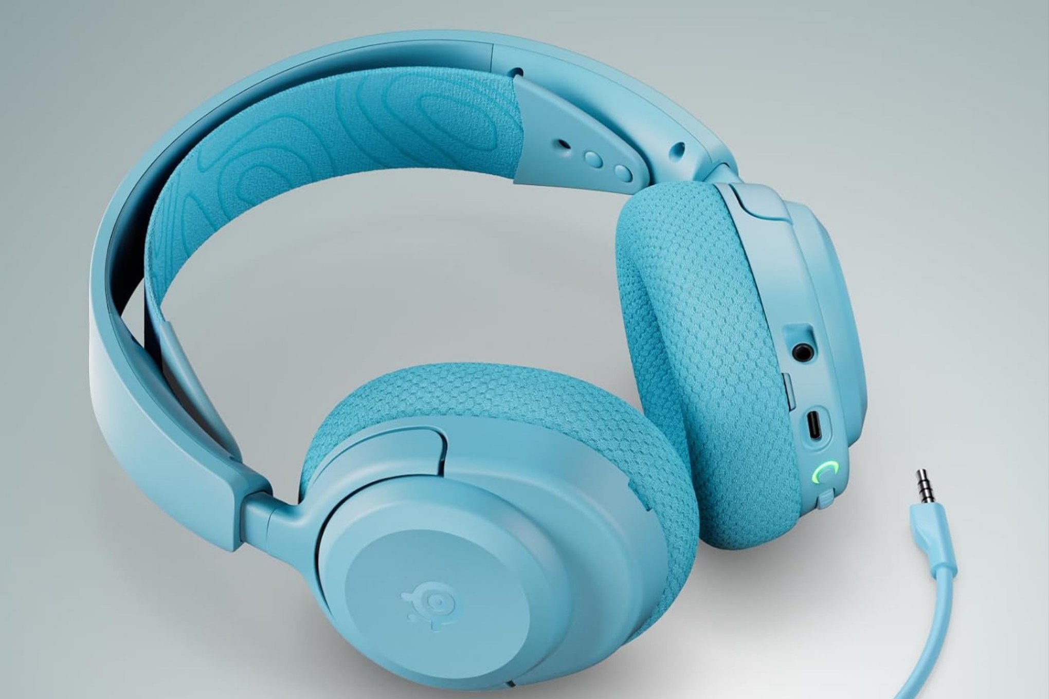 Un par de audífonos inalámbricos de color azul claro con almohadillas de tela texturizada, arco superior acolchado y un cable de 3.5mm conectado en el lateral derecho.