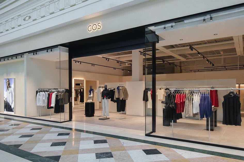 A modern COS retail store with large glass windows and a black frame located in a shopping mall, displaying various clothing items on racks and mannequins inside.