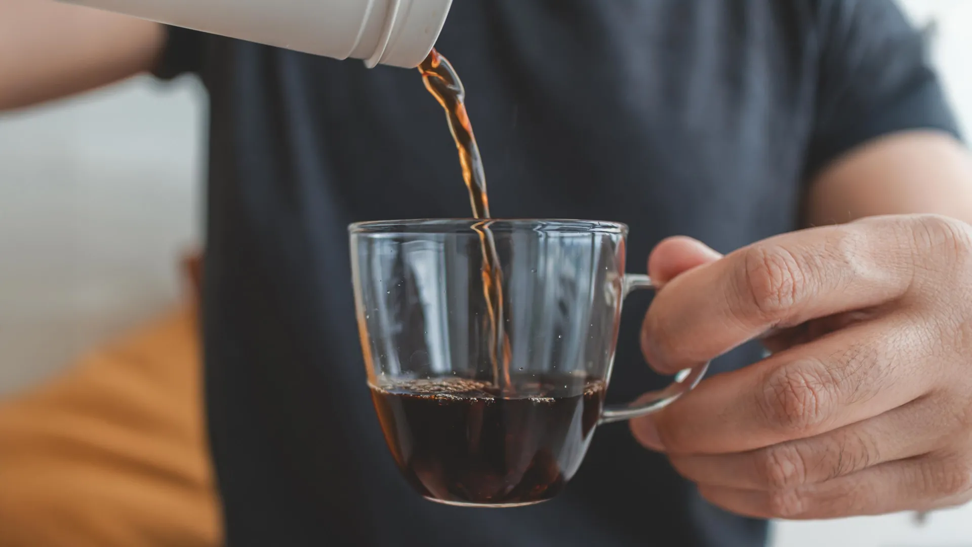 A person pours hot coffee from a drip coffee maker into a glass cup held in hand.