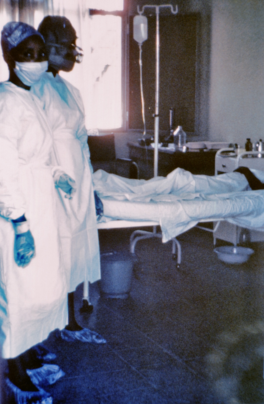 Un trabajador de salud con traje de protección blanco y azul, mascarilla y gafas de protección atiende a un paciente en una cama de hospital con equipos médicos de soporte, incluida una bolsa de suero intravenoso.
