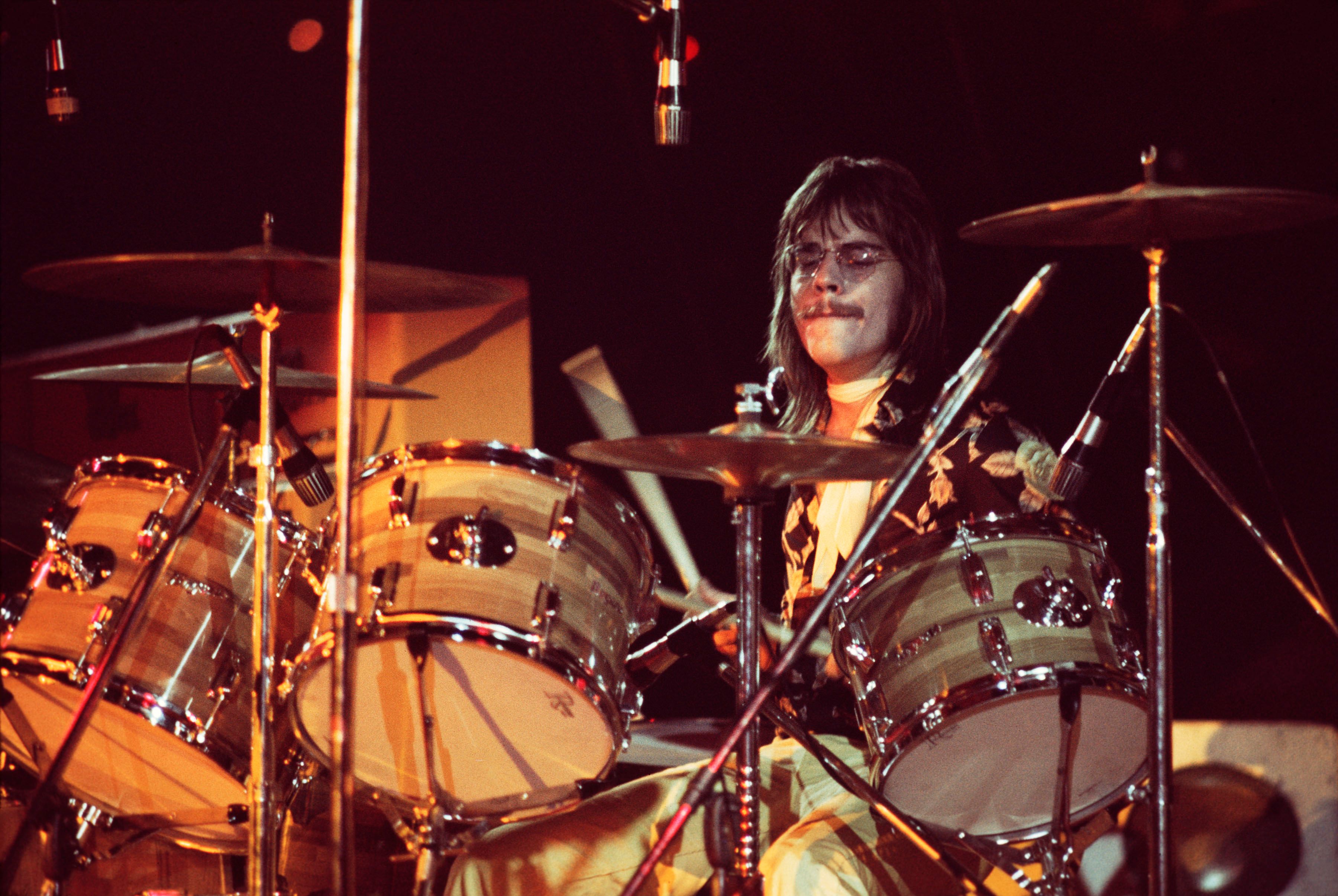 A drummer with long dark hair and glasses plays an expansive drum kit on stage under warm stage lighting during a live performance.