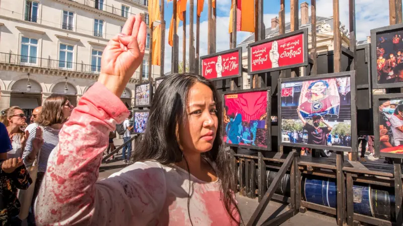 Una mujer con cabello largo y negro levanta el puño en un gesto de protesta en una plaza pública, vistiendo una chaqueta rosa, con pancartas y banderas de Myanmar visibles detrás de ella.