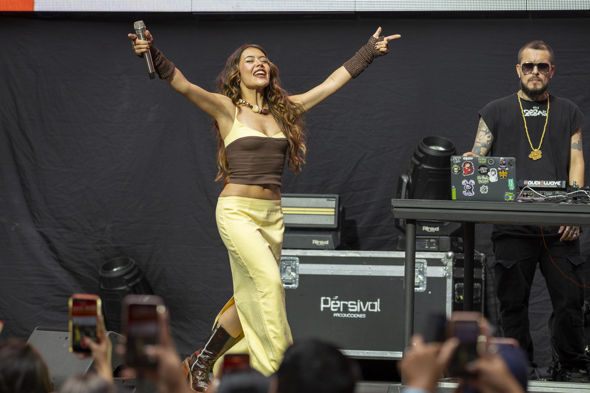 Una cantante con los brazos levantados sosteniendo un micrófono actúa en un escenario mientras un productor musical trabaja en los controles detrás de ella, con el público registrando el evento con sus teléfonos celulares.