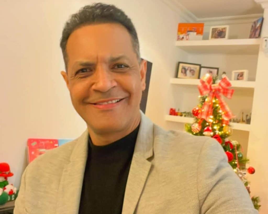 A man smiles at the camera wearing a beige blazer over a black shirt, with red and green Christmas decorations behind him in a room with white shelving.