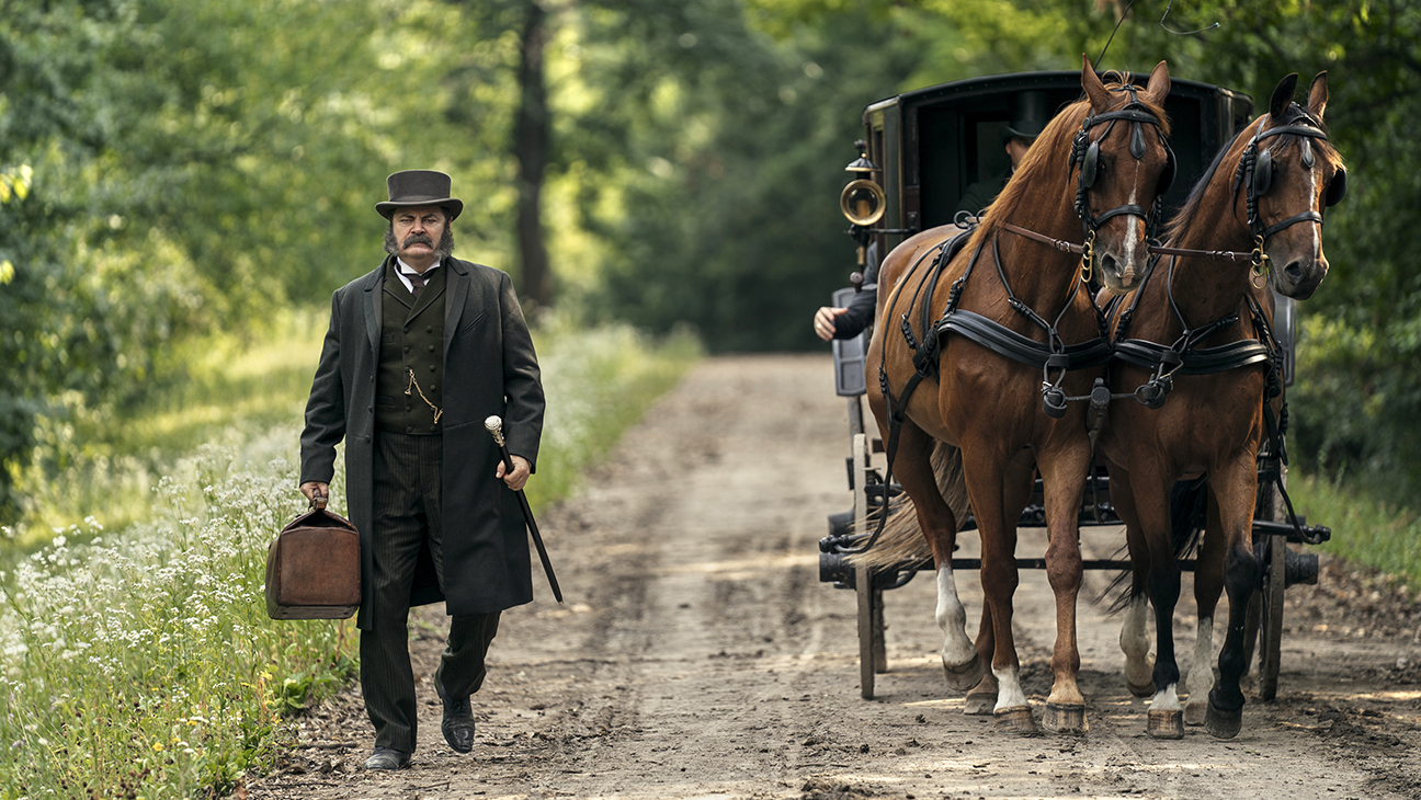 Un hombre vestido con traje oscuro decimonónico y sombrero de copa camina por un camino de tierra sosteniendo una maleta, mientras una carreta tirada por dos caballos se aproxima por detrás.