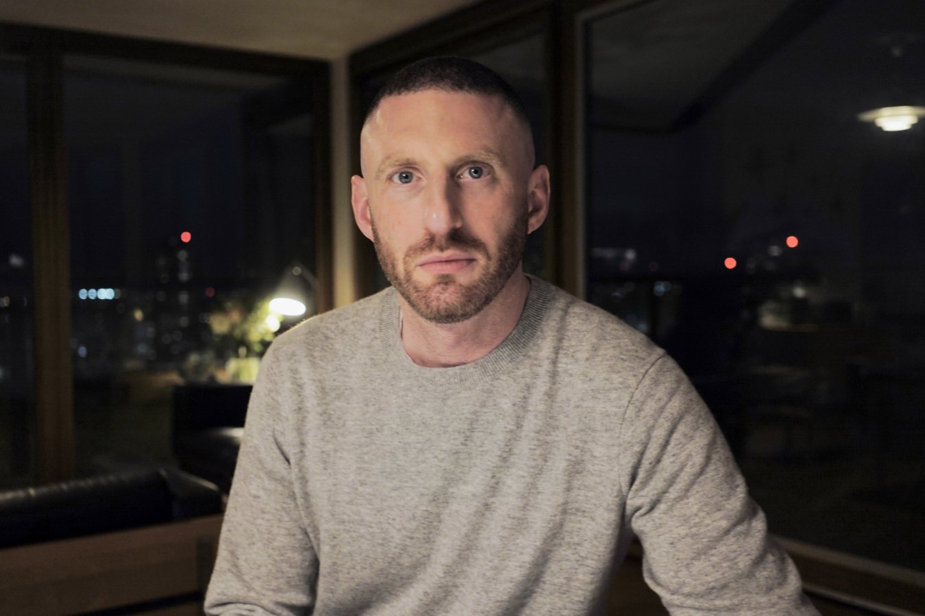A man with a beard wearing a light gray sweater sits indoors in front of large windows showing a nighttime city skyline.