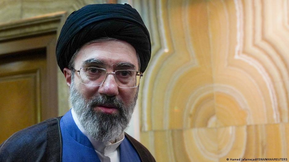 A middle-aged bearded man with grey beard wearing a dark blue suit, black turban and glasses, photographed inside an ornate room with cream-coloured walls and gold decoration.