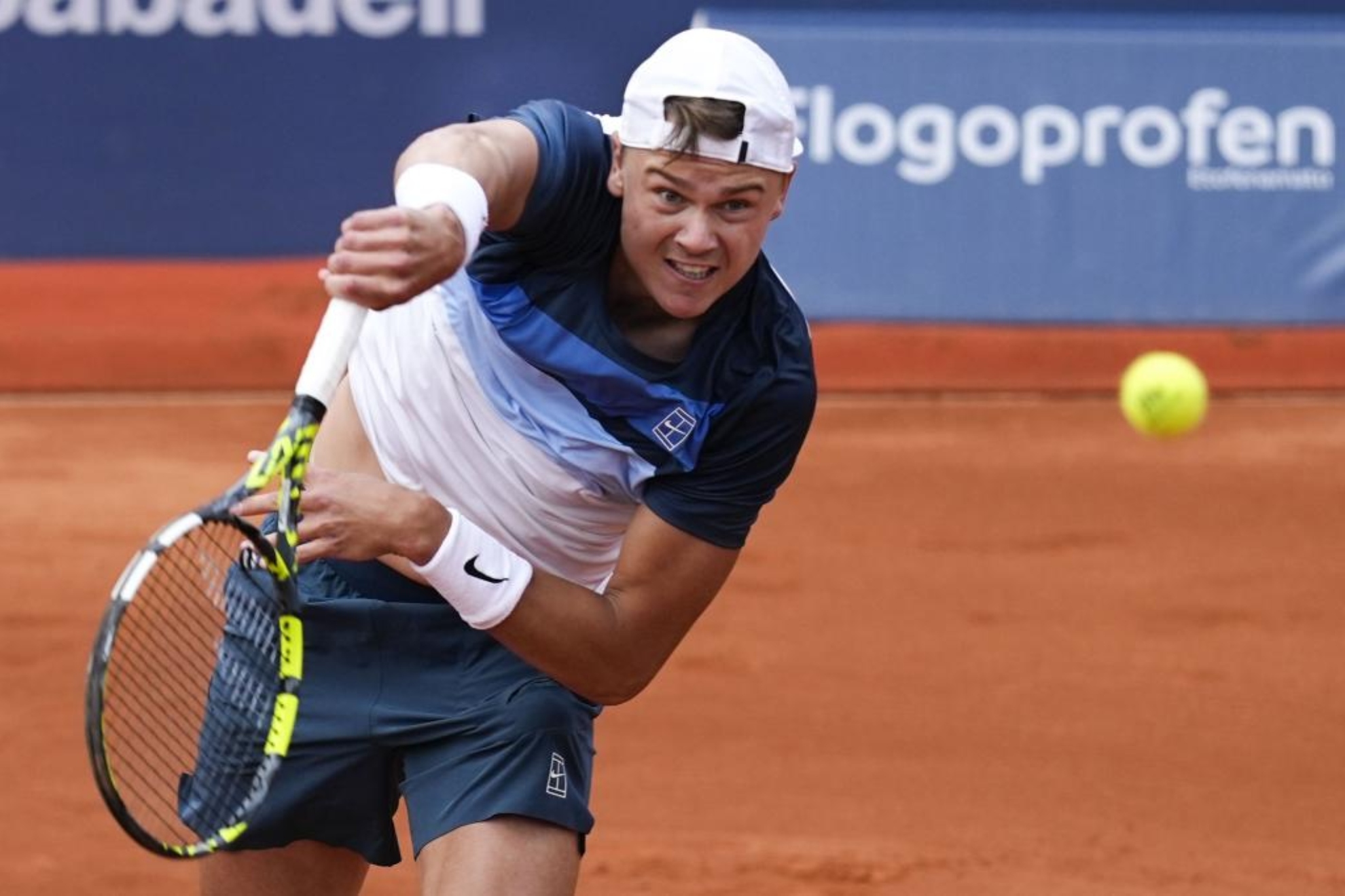 Un tenista con gorra blanca y uniforme azul marino golpea una pelota de tenis en una cancha de arcilla roja durante un partido.