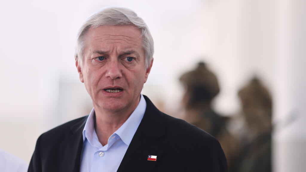 A grey-haired man wearing a black blazer and light blue shirt speaks to the camera with a blurred background showing two people standing.