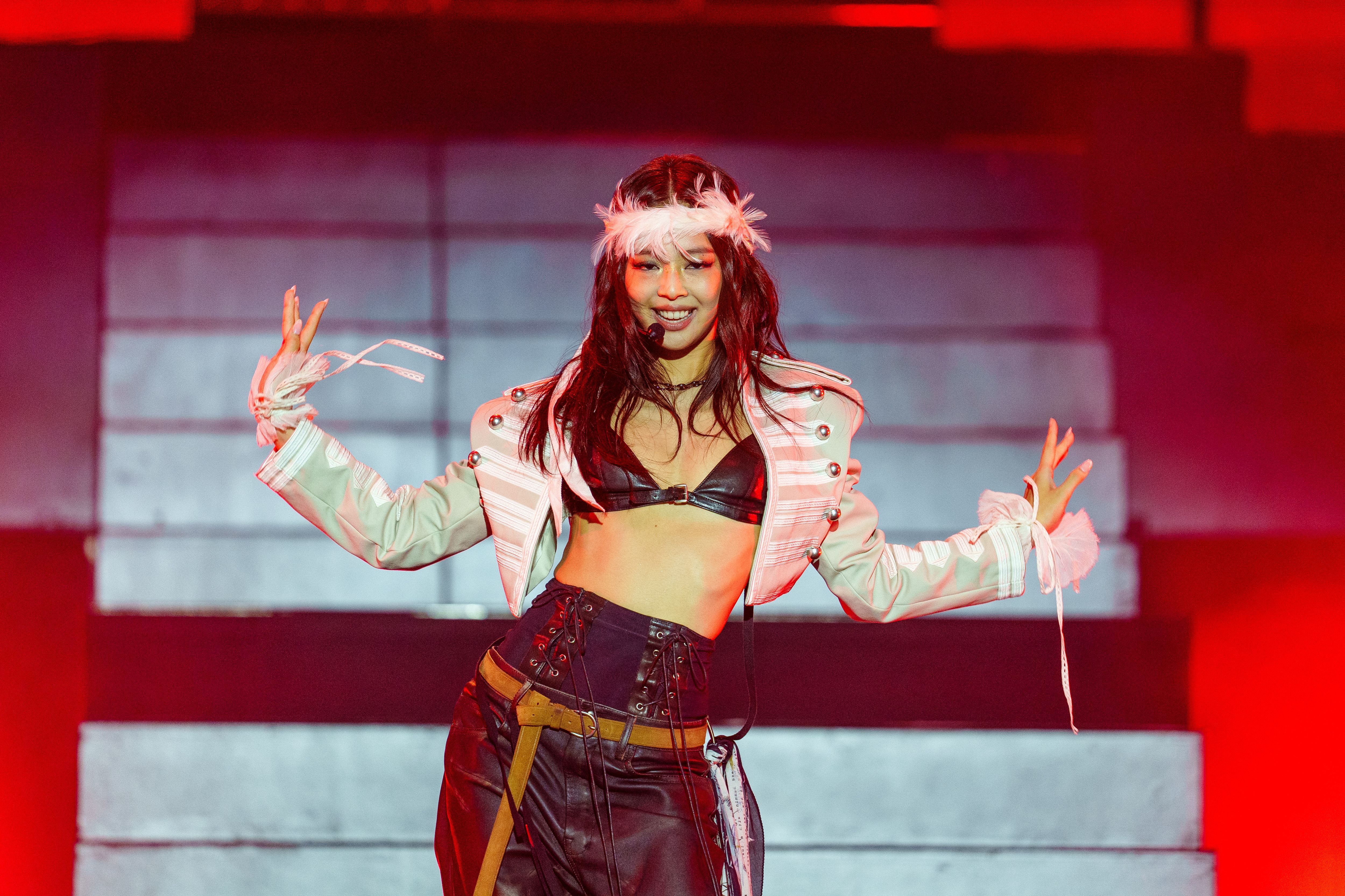 A performer on stage wearing a pink and white striped jacket, black crop top, and dark pants with a metallic belt, with long reddish hair and a feathered headpiece, striking a pose with arms outstretched against a red-tinted background.