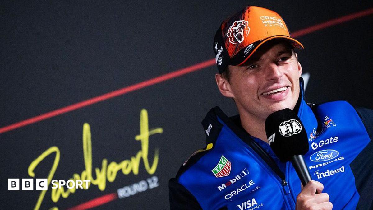 Max Verstappen wearing an orange and red cap and blue team jacket holds a microphone during a press conference, smiling at the camera.