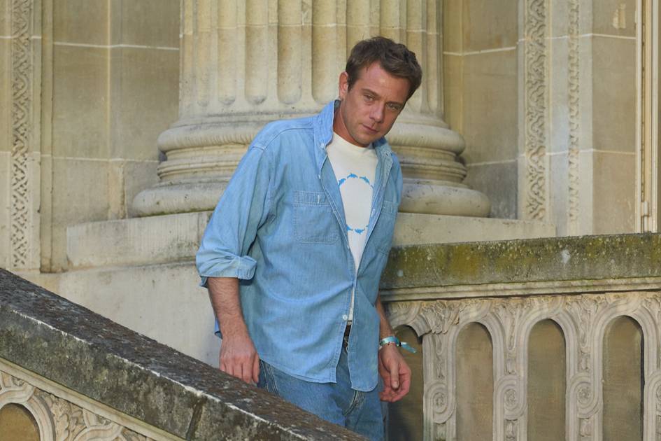 A man in a light blue denim shirt stands against an ornate stone architectural background with classical columns and decorative molding.