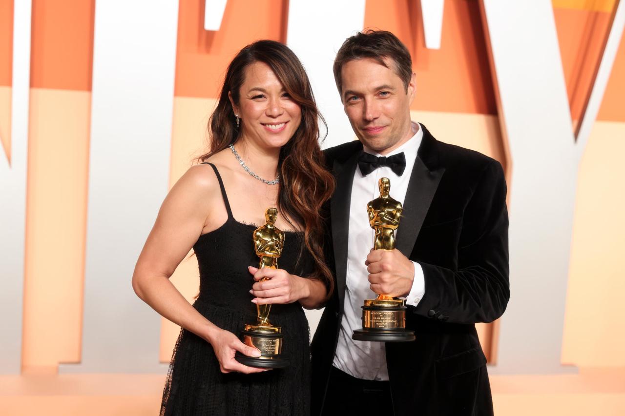Dos personas en traje de gala sostienen estatuillas de Oscar doradas mientras posan frente a un fondo de colores naranja y crema.