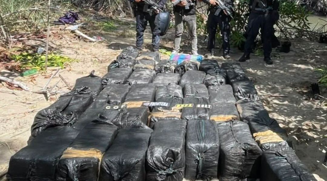 Black packages of confiscated cocaine are arranged on the ground next to uniformed security agents in a mangrove area.