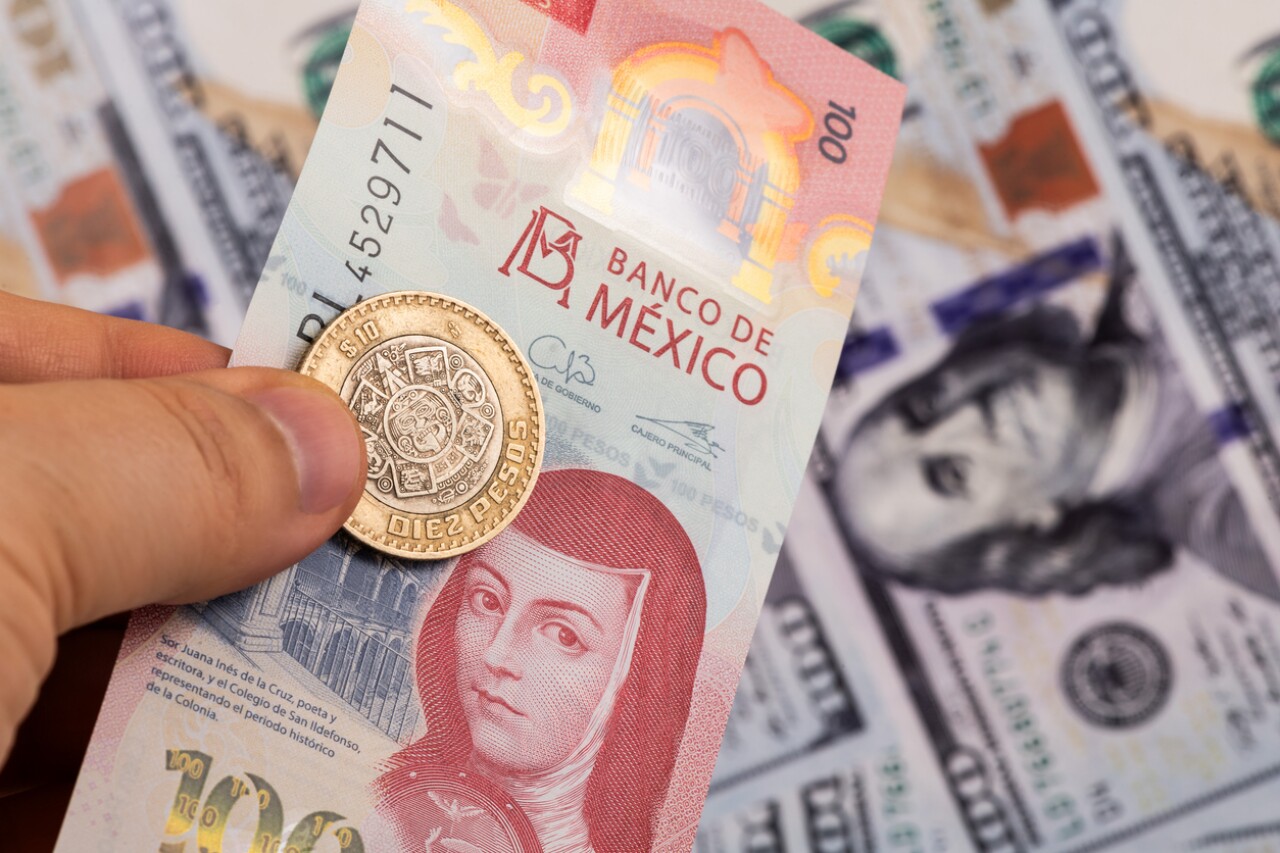 A hand holds a Mexican gold coin over a pink 100-peso note from Banco de México, with banknotes of other currencies blurred in the background.