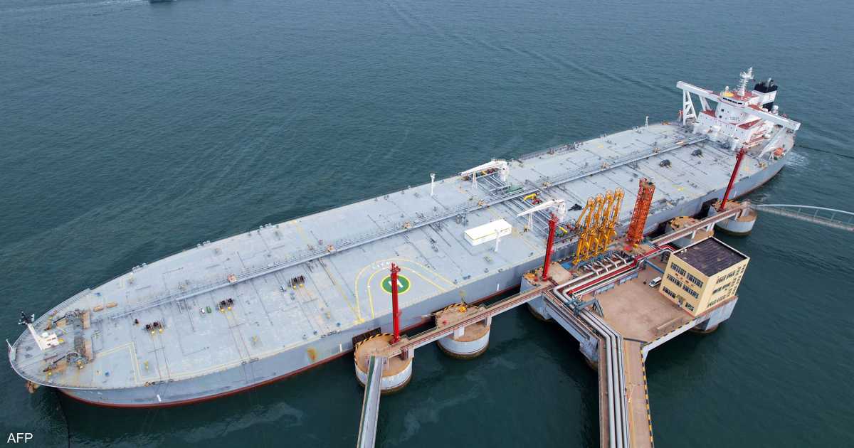 A large white and red tanker vessel docked at a discharge pier with yellow cranes and concrete structure against greyish-blue water.