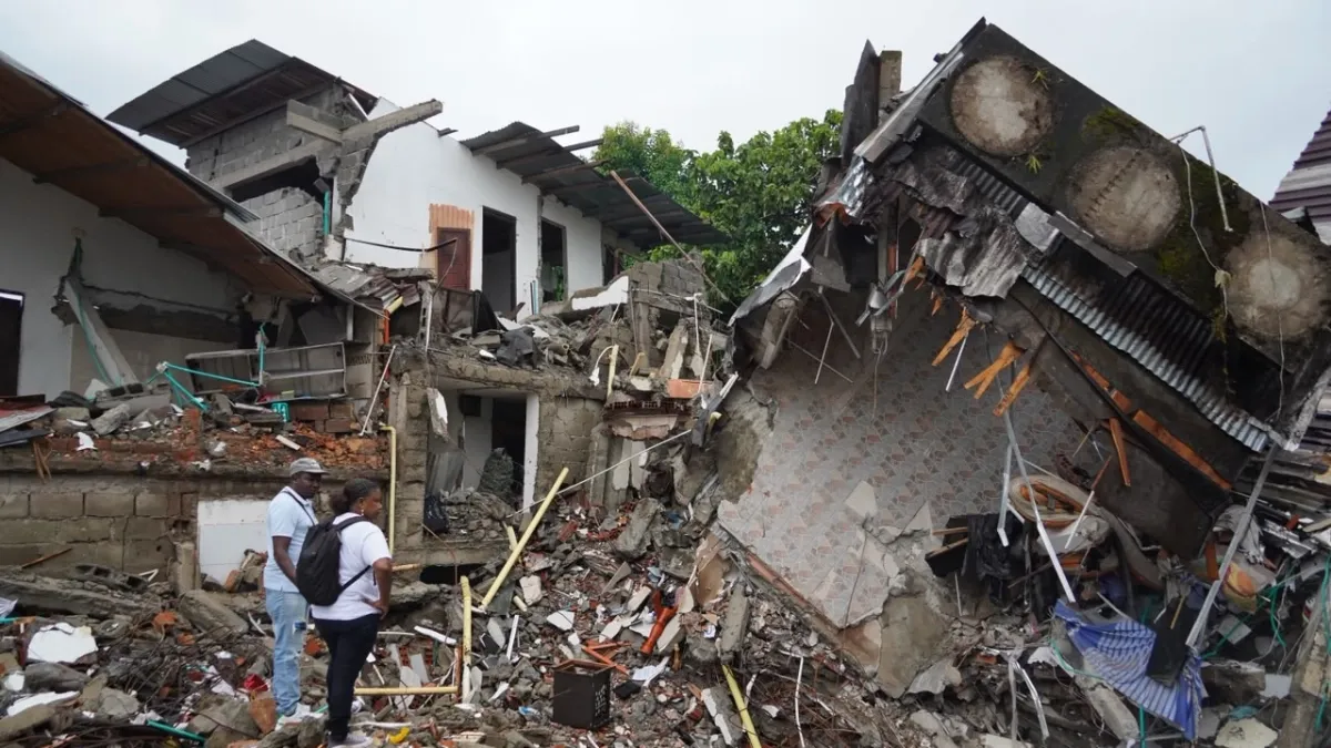 Dos personas de pie observan edificios residenciales destruidos, con escombros, paredes derrumbadas y estructuras de metal expuestas tras un terremoto.